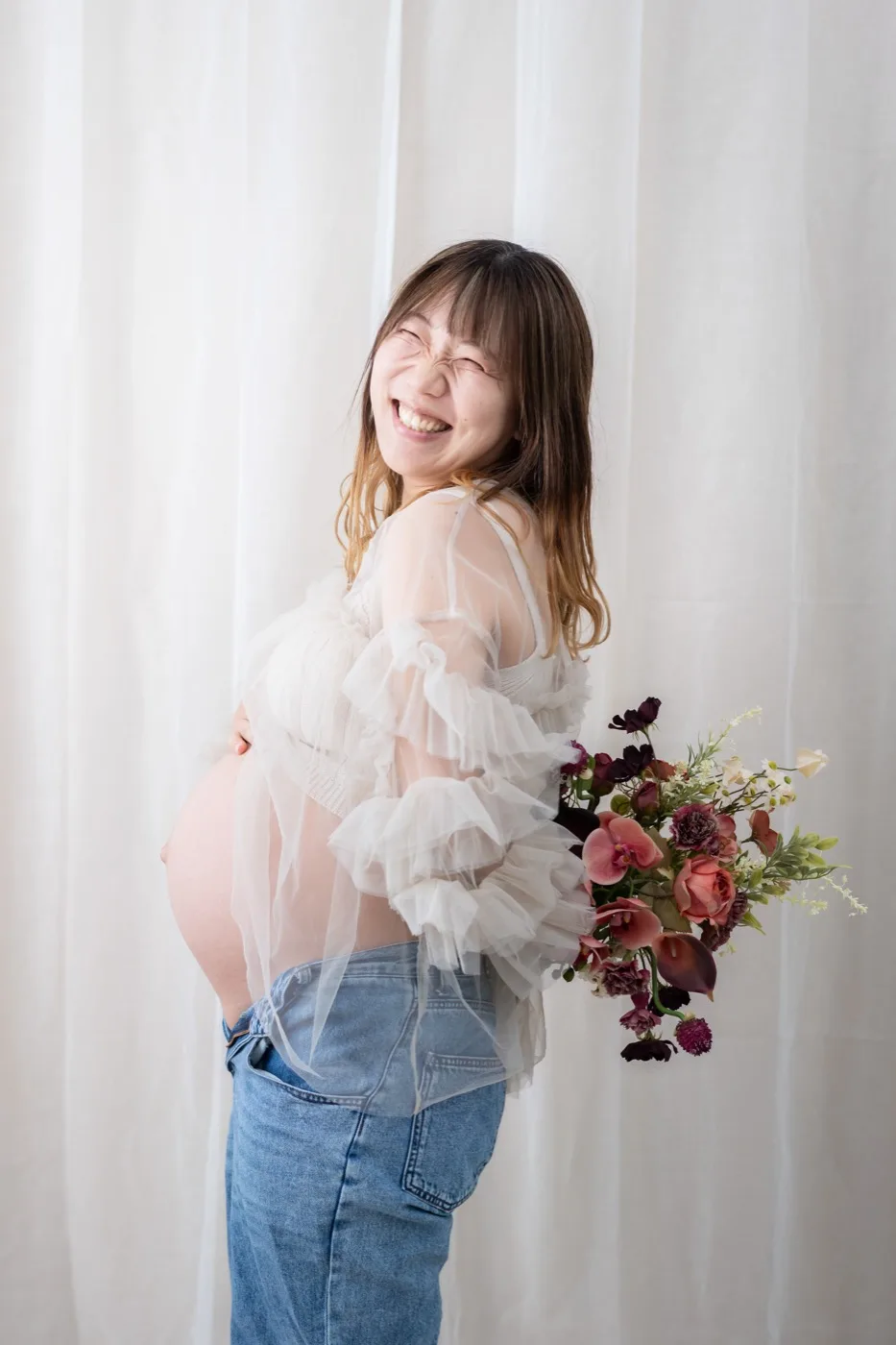 白いカーテンの前で、ブーケを持って笑うチュール衣装の妊婦