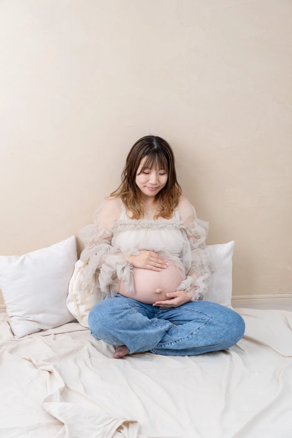 ベッドの上であぐらをかき、おなかに両手を重ねる妊婦