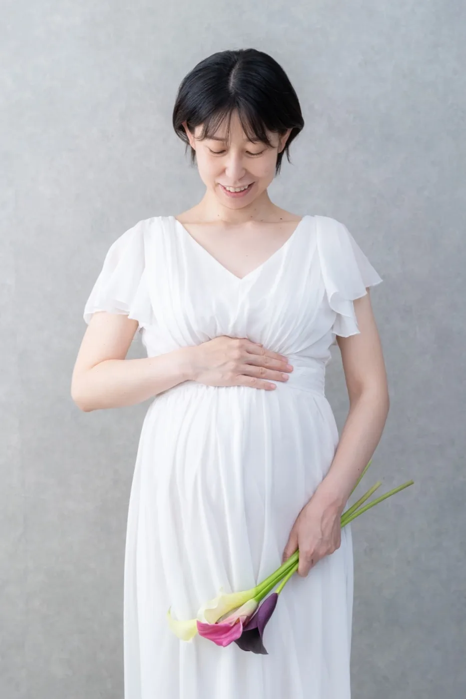 白いドレスでカラーの花を持ち、おなかに手を添える妊婦