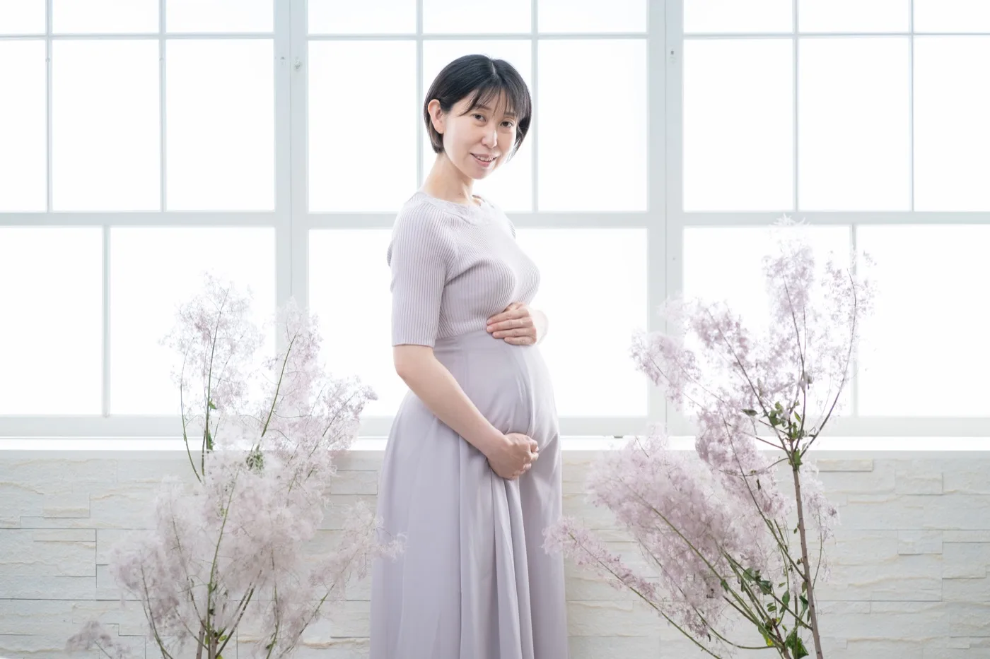 淡い水色のドレスで、スモークツリーに囲まれてこちらを見る妊婦