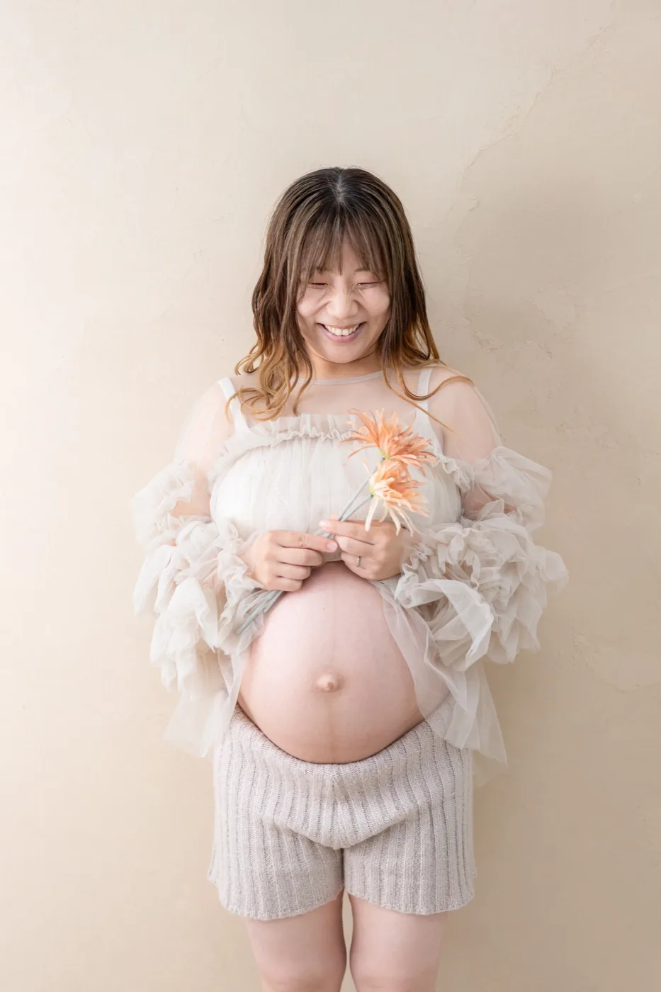 ベージュの壁の前で、ガーベラを持って笑うチュール衣装の妊婦