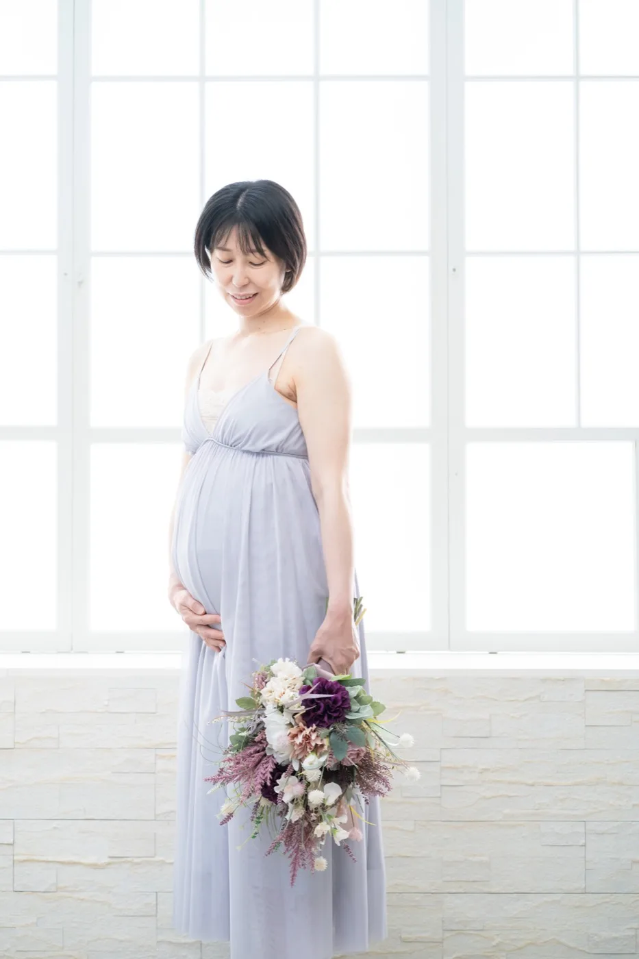 淡い水色のドレスでブーケを持ち、おなかに手を添える妊婦