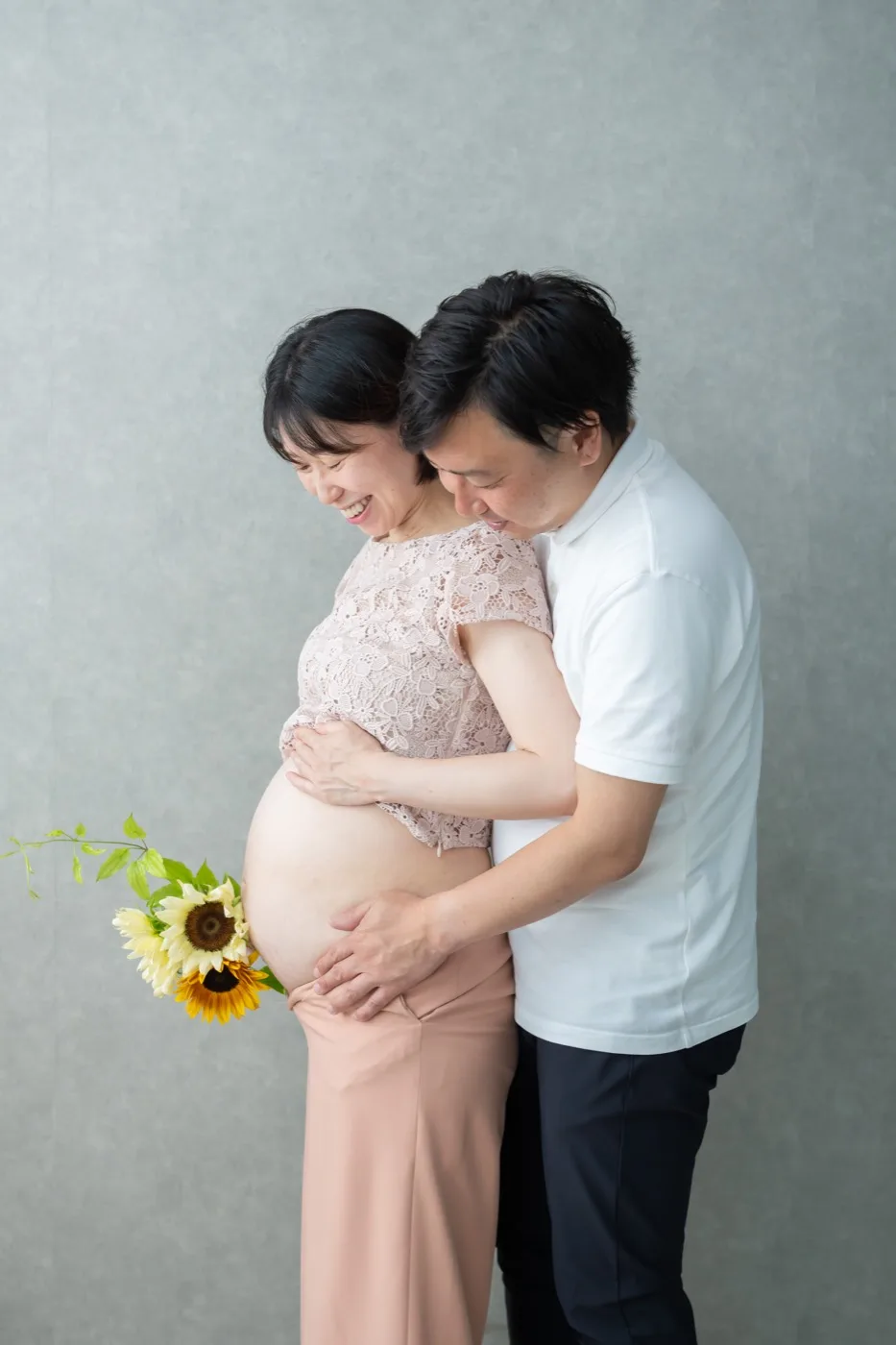 ひまわりを持つ妻のおなかに、後ろから夫が手を添えるマタニティフォト