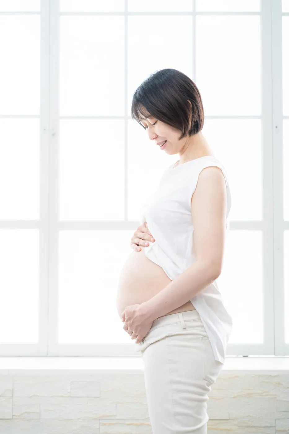 窓辺の光の中で、白いトップスからおなかを出して立つ妊婦