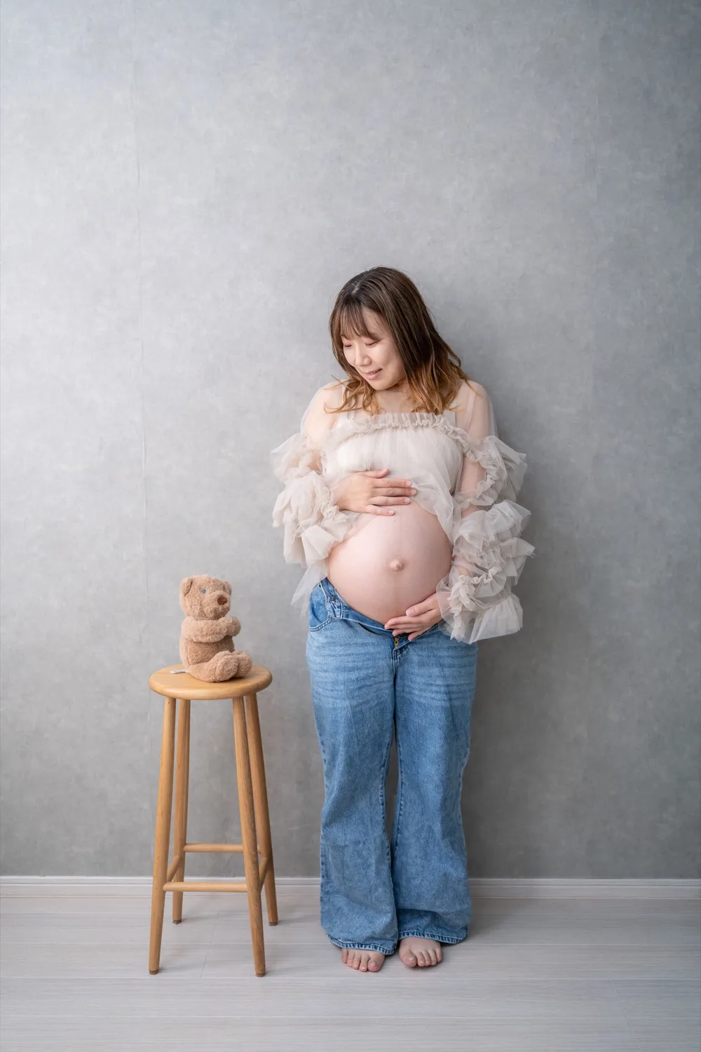 チュールの衣装とデニムで立つ妊婦の全身。かたわらのスツールにくまのぬいぐるみ