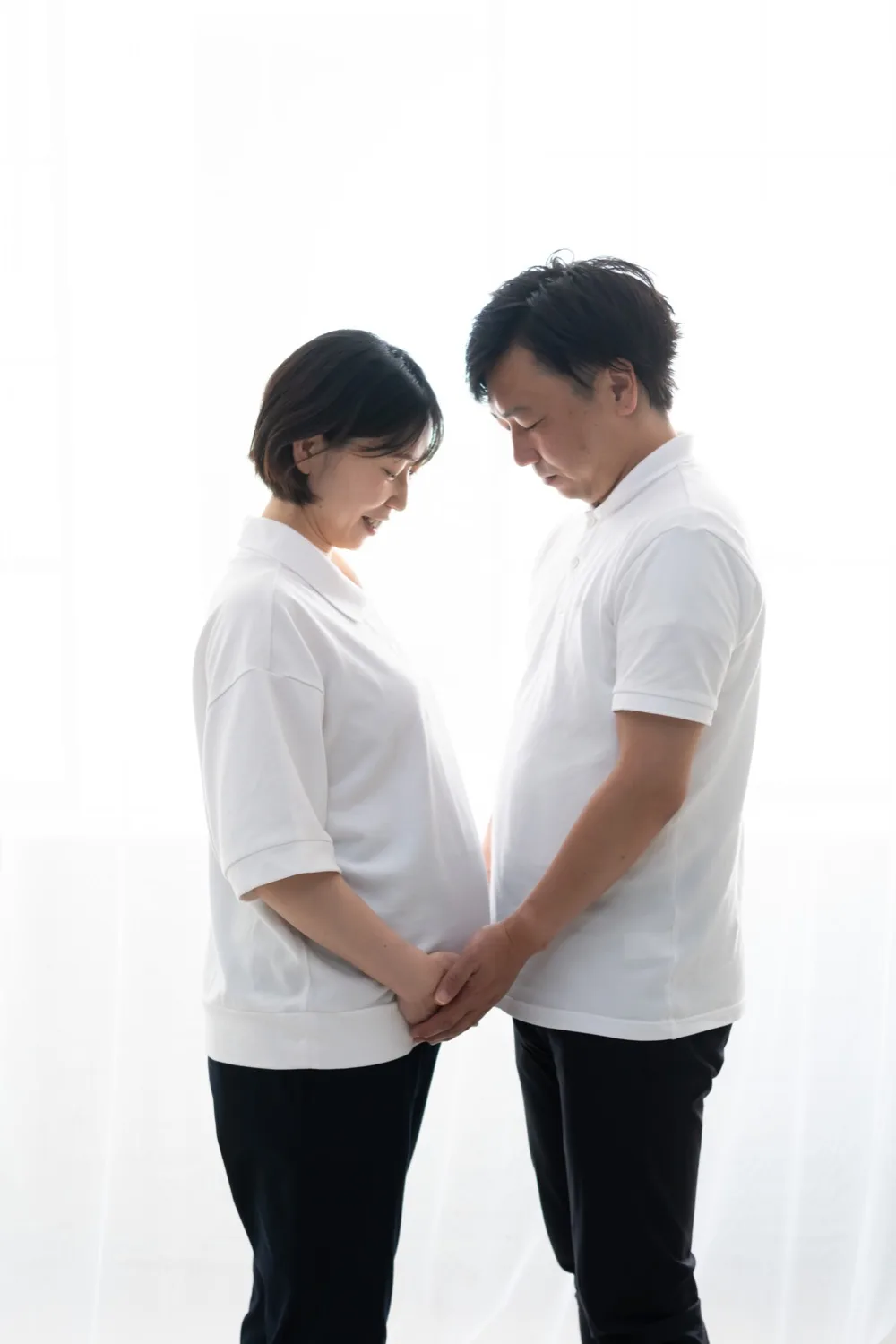 窓辺で向かい合い、おなかの前で手を重ねる夫婦